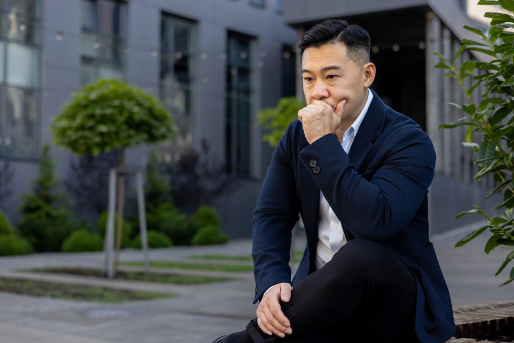 Pensive and serious young Asian male businessman sitting on a bench near an office center and looking upset while holding his head in his hand