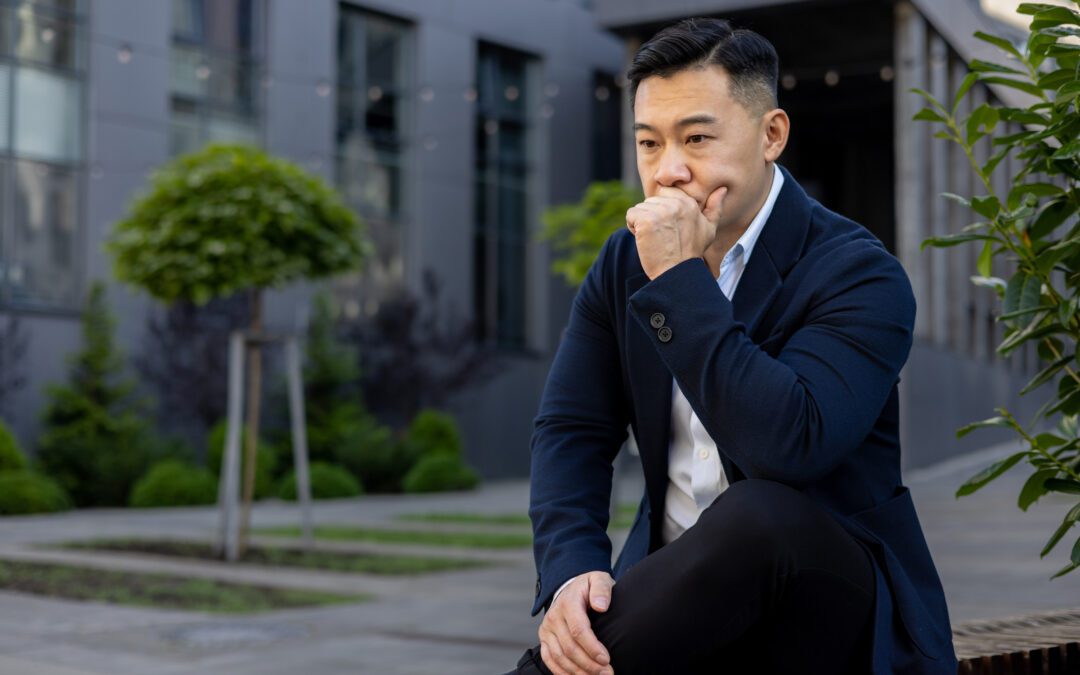 Pensive and serious young Asian male businessman sitting on a bench near an office center and looking upset while holding his head in his hand